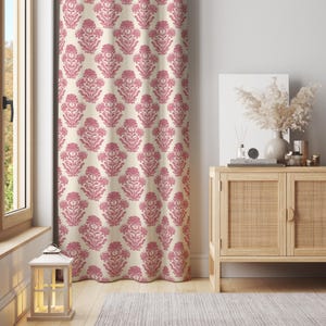 Puede incluir: Una cortina estampada con un diseño floral repetitivo en tonos rosa y crema. La cortina cuelga frente a una ventana, con un mueble de madera y objetos decorativos visibles en el fondo. El patrón floral es simétrico.