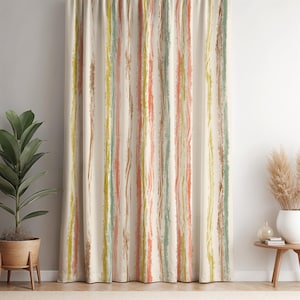 Op de afbeelding: Crèmekleurig gordijn met verticale penseelstreepstrepen in bruin, geel, oranje en groen. Het gordijn hangt voor een witte muur. Een plant in een pot en een kleine tafel met een vaas zijn zichtbaar.