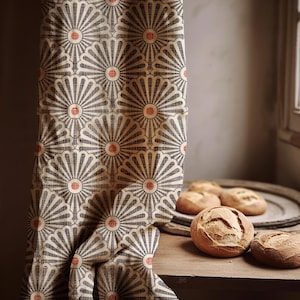 Op de afbeelding: Een gordijn met een herhalend waaiervormig patroon in grijs, beige en rood. Verschillende ronde broden liggen op een houten oppervlak, bij een raam. Het gordijn is gedrapeerd en toont de details van het patroon.