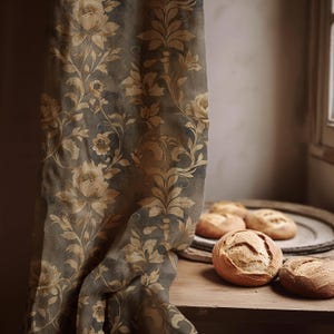 May include: A dark gray fabric curtain featuring a repeating pattern of light tan floral motifs and scrolling vines. The curtain hangs vertically next to a wooden surface holding several round, crusty loaves of bread on a circular plate.