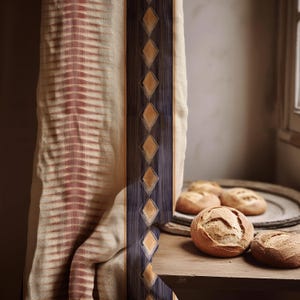 May include: A beige and red patterned curtain with a dark blue and gold diamond design. Several round loaves of bread are on a wooden surface, with more bread on a round tray in the background.