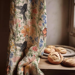 Può includere: Una tenda beige con un motivo floreale con uccelli neri, fiori arancioni, rosa e blu. Diverse pagnotte rotonde sono su un tavolo di legno. La tenda ha un tema botanico.