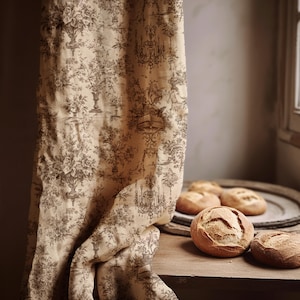 May include: Beige curtain with a brown floral and architectural pattern. Several round loaves of bread sit on a wooden surface next to a decorative metal tray. The curtain hangs near a window.