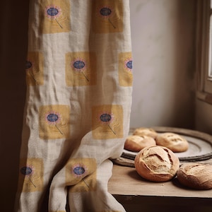 Puede incluir: Un paño de cocina de lino beige con un patrón repetitivo de cuadrados amarillo mostaza, cada uno con un motivo floral bordado en rosa y azul. La tela tiene un tejido texturizado y cuelga verticalmente junto a varios panecillos redondos.