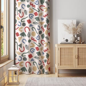May include: Cream-coloured curtain featuring a repeating pattern of colourful abstract shapes, including red squares, blue arches, and green leaf-like forms. The shapes are scattered across the fabric, creating a playful design. The curtain hangs in front of a window.