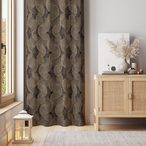 May include: A brown and gold patterned curtain hangs next to a window. The curtain's design features overlapping circles. A wooden cabinet with a vase of dried flowers and candles sits to the right. A white lantern sits on the floor.