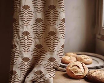 May include: A beige curtain with a repeating brown floral pattern hangs next to a wooden table. Several round loaves of bread sit on the table and a wooden tray. The curtain is draped over the table.