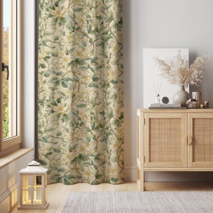 Op de afbeelding: Crèmekleurig bloemengordijn met een patroon van witte bloemen en groene bladeren. Het gordijn hangt bij een raam en een houten kast met decoratieve items.