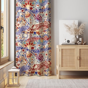 以下が含まれることがあります： クリーム色の背景に、赤、青、黄色の花柄と幾何学模様が繰り返されるデザインの模様入りのカーテン。窓の前にカーテンが吊るされており、背景には木製のキャビネットと装飾品が見えます。