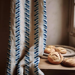 Op de afbeelding: Een linnen gordijn met een blauw patroon hangt naast een houten tafel. Verschillende knapperige broodjes liggen op de tafel en een houten dienblad. Het gordijn heeft een herhalend ontwerp van blauwe penseelstreken.