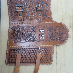 May include: A brown leather holster with floral and geometric tooling. The holster features two buckles, two straps, and the initials "PS" in a circular design. The leather is stitched with a light-colored thread.