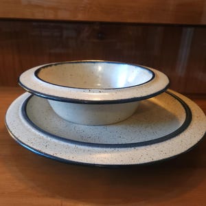 May include: A ceramic bowl and matching plate set featuring a cream-colored base with dark brown speckles throughout. Both pieces have a smooth, rounded design with a distinct dark blue rim accenting the edges of the bowl and the plate.