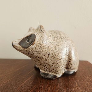May include: A ceramic raccoon figurine featuring a speckled beige and brown glaze. The animal has a dark brown mask around its eyes, pointed ears, and dark brown paws. The sculpture is depicted in a seated position with its head turned slightly.