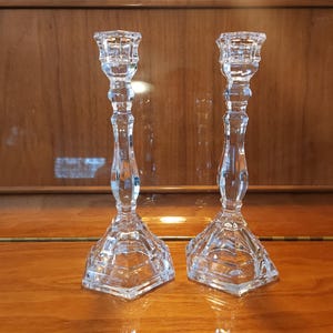 May include: A pair of clear glass candlesticks featuring a faceted, hexagonal base and a slender, multi-tiered stem. Each candlestick has a wide, square-shaped top opening designed to hold a standard taper candle securely in an upright position.