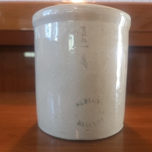 May include: A cylindrical beige stoneware crock with a smooth, glossy finish. The front features the fraction 1/4 printed in black ink near the top rim, and the text ALBERT KLUCLIF printed in black ink near the base of the vessel.