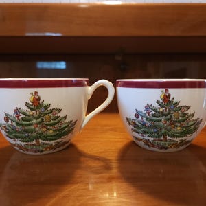 May include: Two white ceramic teacups featuring a festive Christmas tree illustration on the front. Each cup has a dark red band around the top rim and a handle on the right side. The tree is decorated with colorful ornaments and a figure.