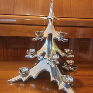 May include: A polished silver-toned metal candle holder shaped like a stylized evergreen tree. The structure features multiple tiered branches, each ending in a small, circular cup designed to hold a candle, creating a symmetrical display.
