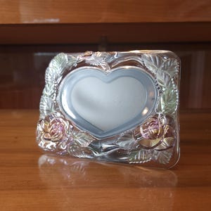 May include: A rectangular clear glass picture frame featuring a central heart-shaped opening. The frame is decorated with molded glass rose blossoms and leaves along the edges, exhibiting an iridescent finish that reflects light in various colors.