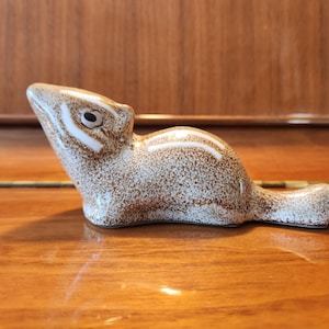 May include: A ceramic chipmunk figurine featuring a speckled brown and white finish. The small animal is depicted in a resting position with a pointed snout, a dark eye, and distinct white stripes running along its head and back.