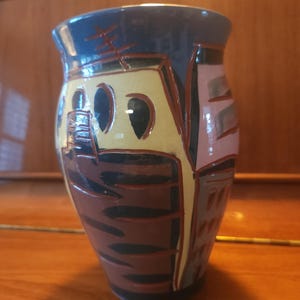 May include: A ceramic vase featuring a glossy finish with a multi-colored abstract design. The surface displays bold blocks of yellow, brown, pink, and blue, separated by dark, incised lines that create a geometric pattern across the exterior.