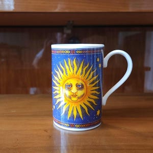 May include: A cylindrical ceramic mug featuring a vibrant yellow sun with a human face centered on a deep blue background filled with small yellow stars. The mug has a white handle and decorative geometric borders at the top and bottom edges.