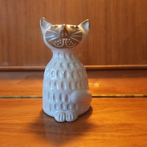 May include: A ceramic cat figurine featuring a cream-colored body with a textured pattern of vertical indentations. The cat has a smooth, light brown face with etched details for the eyes, nose, whiskers, and mouth, and pointed ears.