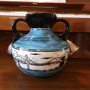 May include: A ceramic vase with two black handles and a glossy black rim. The body features a hand-painted desert landscape in black on a white band, set against a bright blue sky and base. White feathers are attached to the handles with black beads.