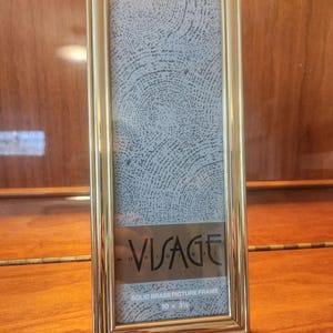 May include: A tall, rectangular solid brass picture frame with a polished gold-tone finish. The frame contains a decorative insert featuring a gray circular pattern and the text "VISAGE ORIGINAL SOLID BRASS PICTURE FRAME 10 x 3.5 inches".