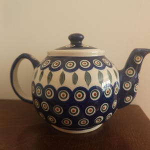 May include: A ceramic teapot featuring a rounded body and a matching lid. The surface is decorated with a repeating pattern of blue, green, and orange circular motifs, interspersed with green leaf-shaped designs on a white background.