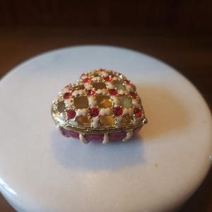 May include: A heart-shaped trinket box featuring a gold-tone metal lattice design accented with small red rhinestones. The top of the box has a cream-colored enamel finish, while the base is a solid deep red color with a smooth, glossy surface.