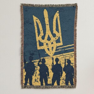 May include: A woven throw blanket featuring the Ukrainian coat of arms in yellow against a blue background. Silhouetted figures of soldiers stand below the emblem. The blanket has a fringed edge.