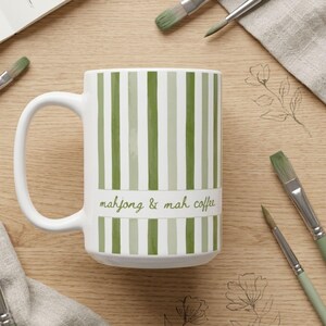 Puede incluir: Una taza de cer&aacute;mica blanca con rayas verticales en varios tonos de verde. Una banda blanca horizontal en el centro muestra el texto "mahjong & mah coffee" en una tipograf&iacute;a cursiva verde. La taza tiene un asa curva en el lado izquierdo.