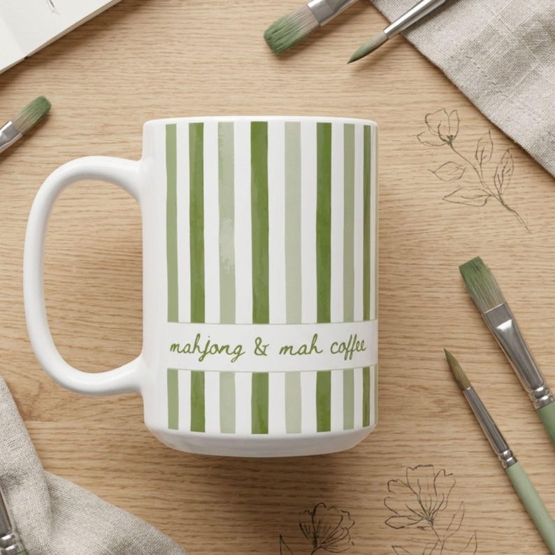 Puede incluir: Una taza de cer&aacute;mica blanca con rayas verticales en varios tonos de verde. Una banda blanca horizontal en el centro muestra el texto "mahjong & mah coffee" en una tipograf&iacute;a cursiva verde. La taza tiene un asa curva en el lado izquierdo.