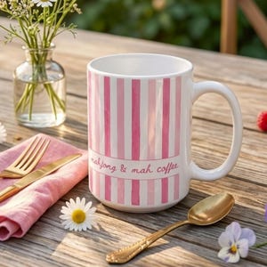 Puede incluir: Una taza de cer&aacute;mica blanca con rayas verticales de color rosa y rosa claro. Una banda blanca horizontal en el centro muestra el texto mahjong & mah coffee en una fuente cursiva rosa. La taza tiene un asa curva y un acabado liso.