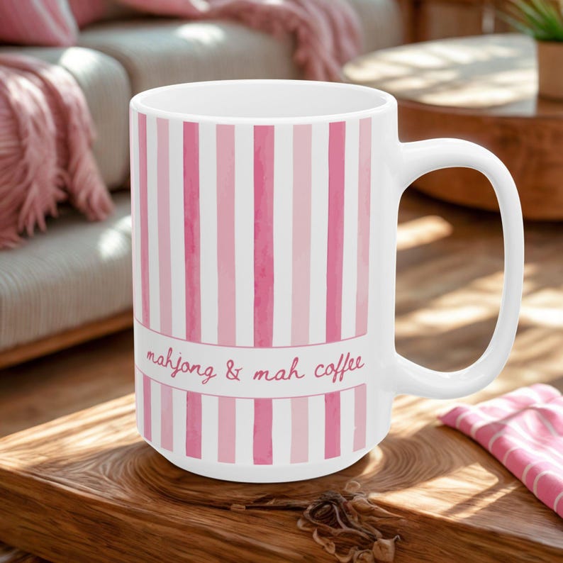 Puede incluir: Una taza de cer&aacute;mica blanca con rayas verticales en varios tonos de rosa. Una banda horizontal en el centro contiene el texto "mahjong & mah coffee" escrito en una fuente cursiva de color rosa. La taza tiene un asa curva a la derecha.