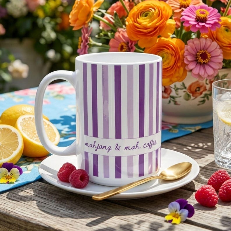 Puede incluir: Una taza de cer&aacute;mica blanca con rayas verticales moradas y lavanda, que presenta el texto "mahjong & mah coffee" en letra cursiva. La taza descansa sobre un plato blanco a juego con una cuchara dorada, rodeada de frambuesas frescas y limones.