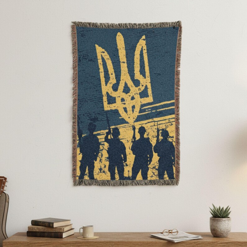 May include: A woven throw blanket featuring the Ukrainian coat of arms in yellow against a blue background. Silhouetted figures of soldiers stand below the emblem. The blanket has a fringed edge.