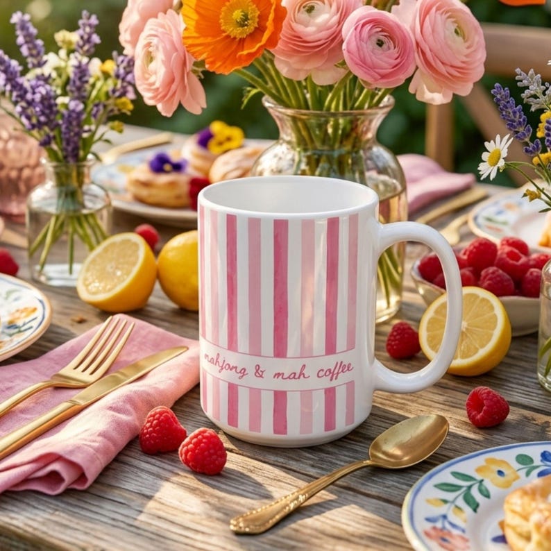 Puede incluir: Una taza de cer&aacute;mica blanca con rayas verticales rosas y el texto "mahjong & mah coffee" impreso en letra cursiva rosa. La taza tiene un asa curva y se encuentra sobre una mesa de madera rodeada de frambuesas frescas, limones y flores.
