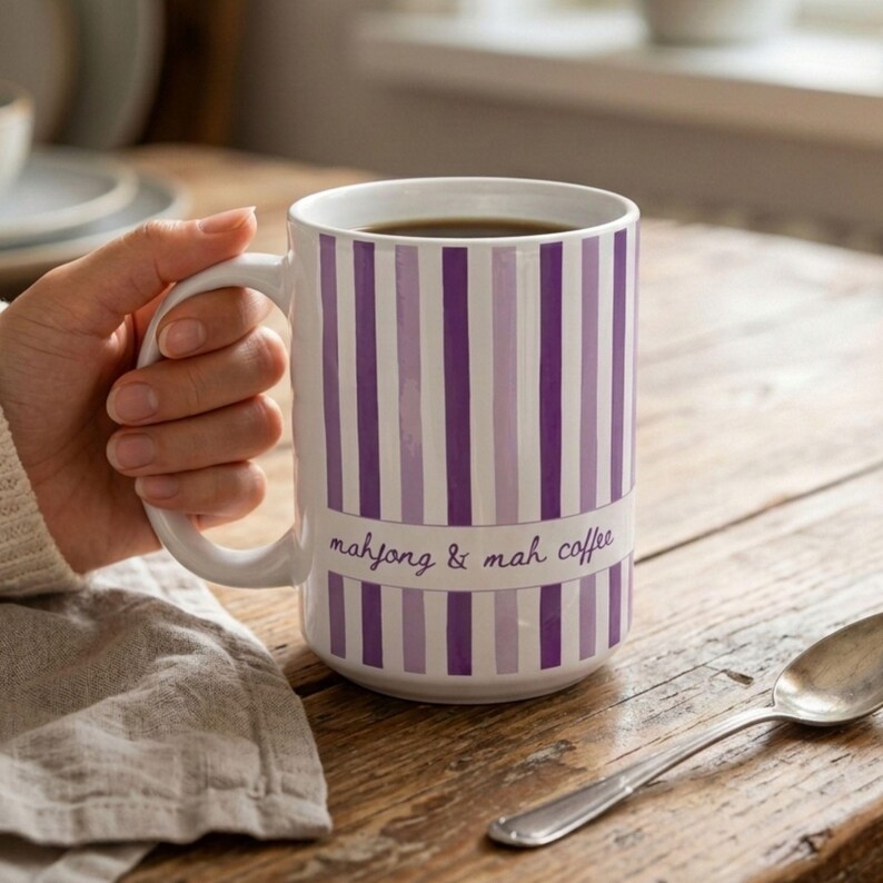 Puede incluir: Una taza de cer&aacute;mica blanca con rayas verticales en varios tonos de morado. Una banda horizontal en el centro contiene el texto "mahjong & mah coffee" en una fuente cursiva morada. La taza tiene un asa curva y un cuerpo cil&iacute;ndrico.