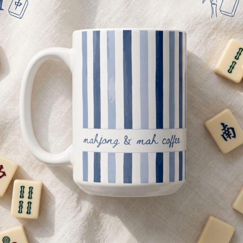Puede incluir: Una taza de cer&aacute;mica blanca con rayas verticales en varios tonos de azul. Una banda horizontal en el centro contiene el texto "mahjong & mah coffee" en una fuente cursiva azul. La taza tiene un asa curva en el lado izquierdo.