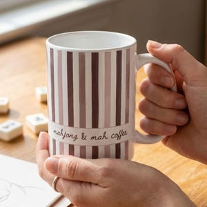 Puede incluir: Una taza de caf&eacute; de cer&aacute;mica blanca con rayas verticales en tonos marr&oacute;n y malva. Una banda horizontal en el centro muestra el texto "mahjong & mah coffee" en una tipograf&iacute;a cursiva de color marr&oacute;n oscuro. La taza tiene un asa curva.