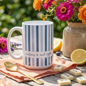Puede incluir: Una taza de cer&aacute;mica blanca con rayas azules verticales y el texto mahjong & mah coffee impreso en letra azul. La taza est&aacute; sobre una servilleta color melocot&oacute;n junto a una cuchara dorada y varias fichas de mahjong sobre una superficie de madera.