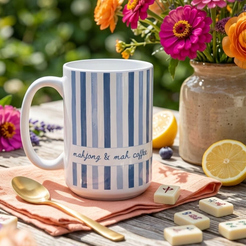 Puede incluir: Una taza de cer&aacute;mica blanca con rayas azules verticales y el texto mahjong & mah coffee impreso en letra azul. La taza est&aacute; sobre una servilleta color melocot&oacute;n junto a una cuchara dorada y varias fichas de mahjong sobre una superficie de madera.