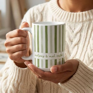 Puede incluir: Una taza de cer&aacute;mica blanca con rayas verticales en varios tonos de verde. Una banda horizontal en el centro muestra el texto mahjong & mah coffee en una fuente cursiva verde. La taza tiene un asa curva en el lado izquierdo.