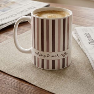 Puede incluir: Una taza de cer&aacute;mica blanca llena de caf&eacute;, con rayas verticales en tonos malva y marr&oacute;n oscuro. Una banda horizontal en el centro muestra el texto en cursiva mahjong & mah coffee. La taza tiene un asa curva en el lado izquierdo.