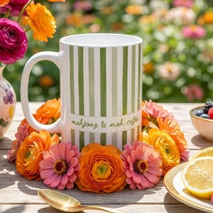 Puede incluir: Una taza de cer&aacute;mica blanca con rayas verticales verdes y el texto mahjong & mah coffee impreso en verde. La taza est&aacute; rodeada por un anillo de flores naranjas y rosas vibrantes, con una cuchara dorada sobre la superficie de madera.
