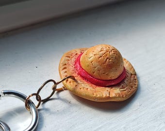May include: A miniature straw hat keychain with a red band. The hat is a light tan color with a textured surface and is attached to a silver keyring with gold-colored chain links. The keychain is resting on a white surface.