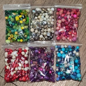 May include: Six clear plastic bags filled with various beads in different colors. The bags contain beads in shades of green, white, pink, red, purple, and blue. The beads vary in size and shape, including pearls and faceted beads.