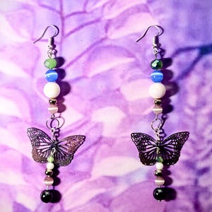 May include: Dangle earrings featuring a butterfly design. The earrings have a silver hook, with a series of beads in green, blue, white, and pink. A detailed, openwork butterfly charm hangs below, with a black and dark brown finish.