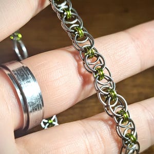 May include: A silver chainmail bracelet with green accents. The bracelet is made of small, interlocking metal rings, with green rings interspersed throughout. A silver ring is visible in the background.